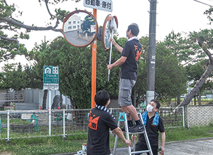 脚立に立ちカーブミラーをモップで拭いている男性と、脚立を左右から押さえている男性の写真