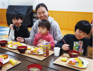 二人の子ども、女性とその膝に座っている子どもの4人が笑顔で食事をしている写真
