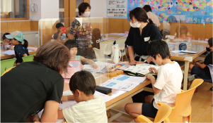 昨年のみんなで宿題3day’sの様子。テーブルで勉強をしている子どもたちとそれを見守る自治協委員たちの写真