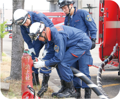 ホースを繋いだ消火栓を紺色のユニホームにヘルメットを着用した３人の隊員が覗き込んでいる写真