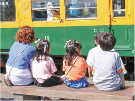 女性と女の子２人、男の子１人がかぼちゃ電車をベンチに座りながら眺めている写真