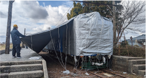 プラットホームからかぼちゃ電車に掛けたシルバーシートをヘルメットに雨具のかぼちゃ電車保存会メンバーが引いている写真